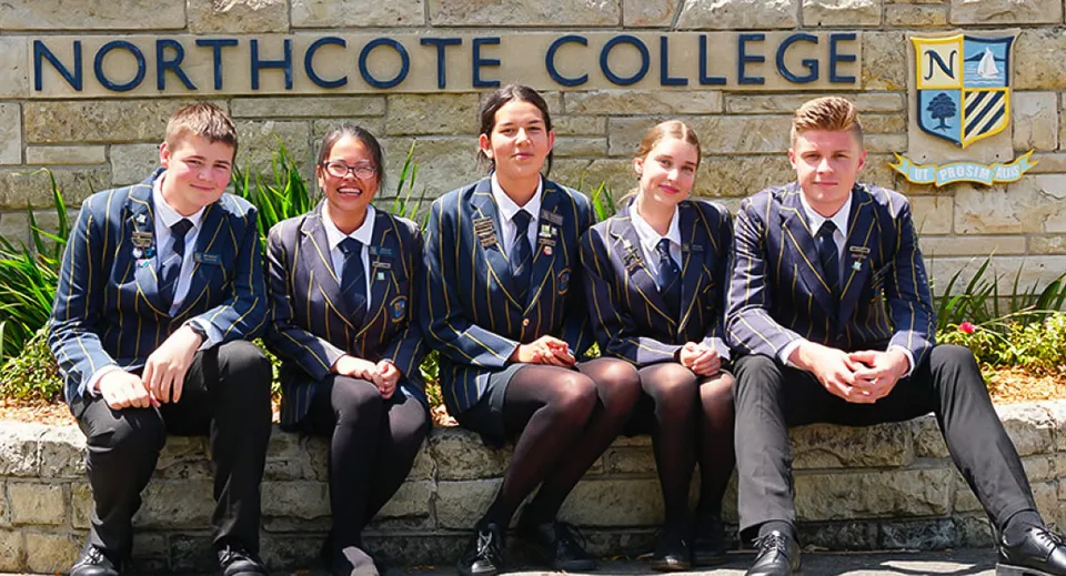 Un gruppo di cinque studenti in divisa scolastica siede su una panchina davanti a un cartello del Northcote College.
