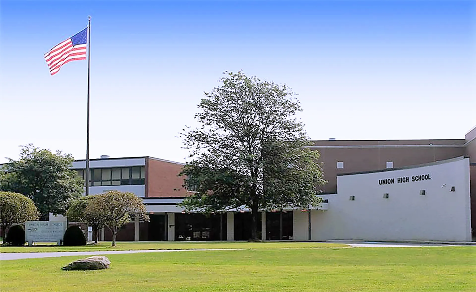 Una grande bandiera americana sventola nel campus della Union High School davanti a un edificio in mattoni e vegetazione verde.