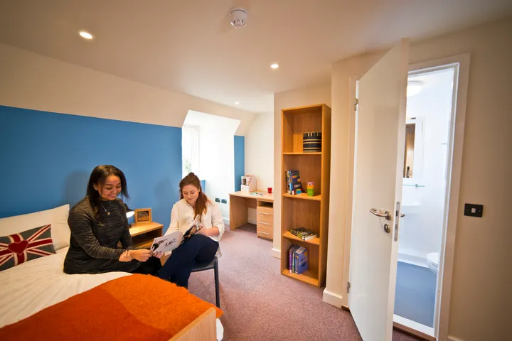 Habitación de la residencia con pared azul, dos estudiantes sentadas en la cama y baño privado al lado