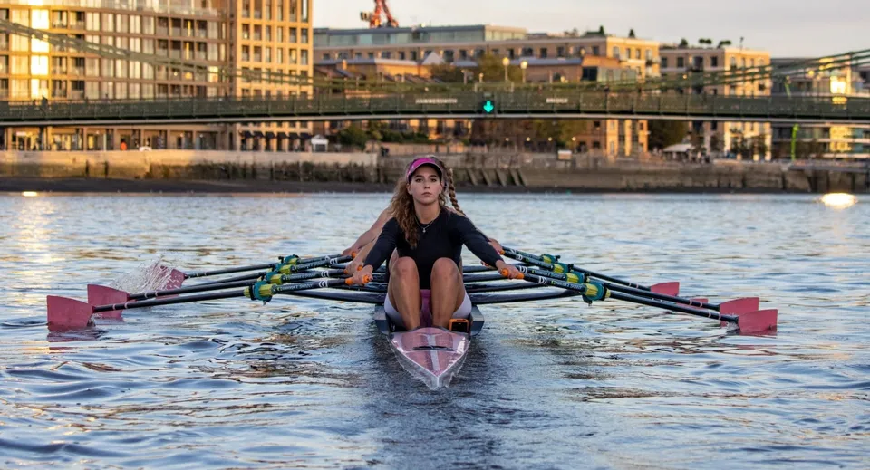 Una persona rema su una barca rosa sull'acqua con lo sfondo della Westminster Secondary School al tramonto.