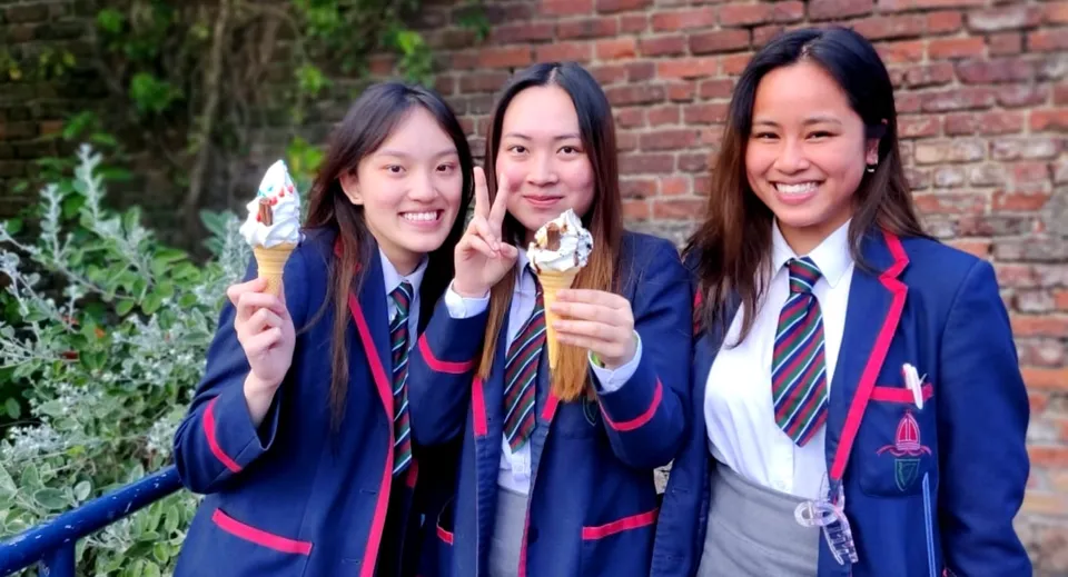 Tre studentesse della Royal School indossano la divisa scolastica e tengono un gelato in un giardino con muro di mattoni.