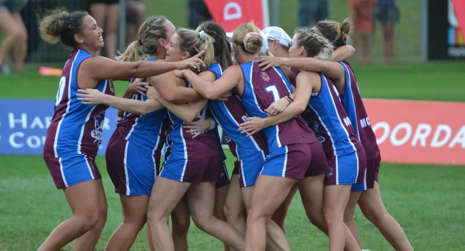 Un gruppo di atlete della Palm Beach Currumbin State High School si abbraccia sul campo sportivo davanti a una bandiera bianca e rossa.