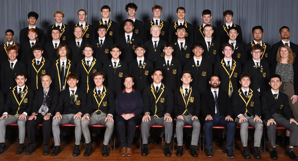 Studenti della Whangarei Boys’ High School seduti in abbigliamento accademico formale in file davanti a uno sfondo grigio.