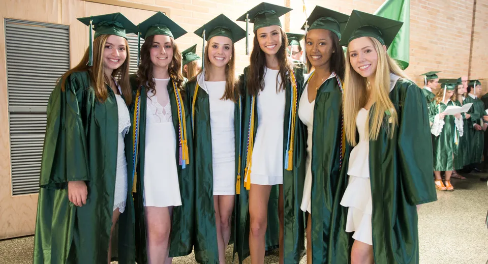 Un gruppo di giovani donne in toga da diploma verde festeggia il proprio diploma davanti a una parete nel campus della Cavendish Road State High School.