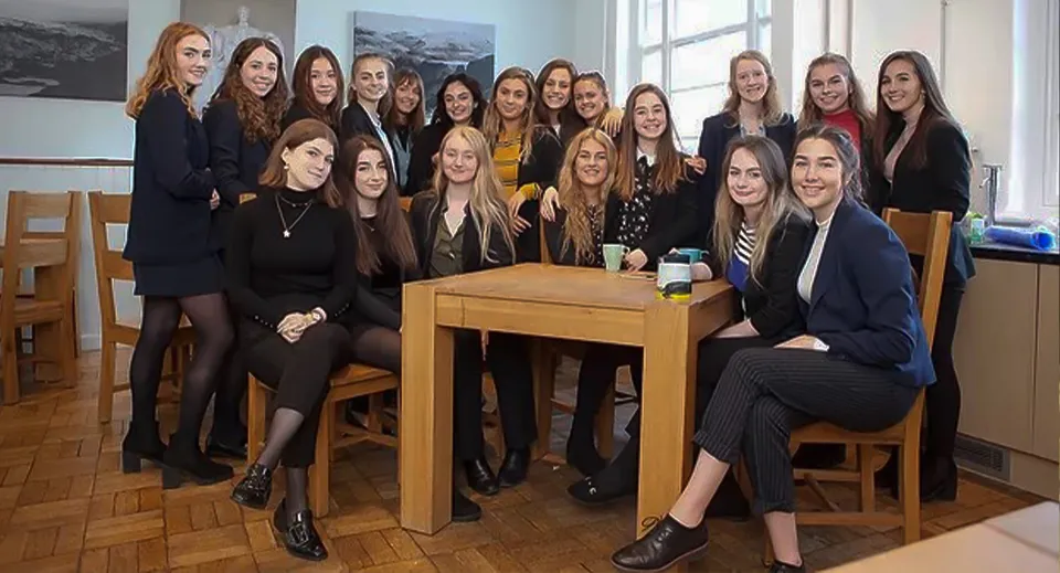 Un gruppo di giovani donne del King William's College siede in abito formale attorno a un tavolo in un'aula con vista sul paesaggio.