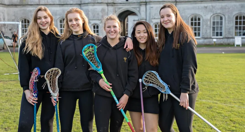 Un gruppo di studentesse della Milton Abbey School è in piedi con le mazze da lacrosse davanti a un edificio storico con finestre ad arco.