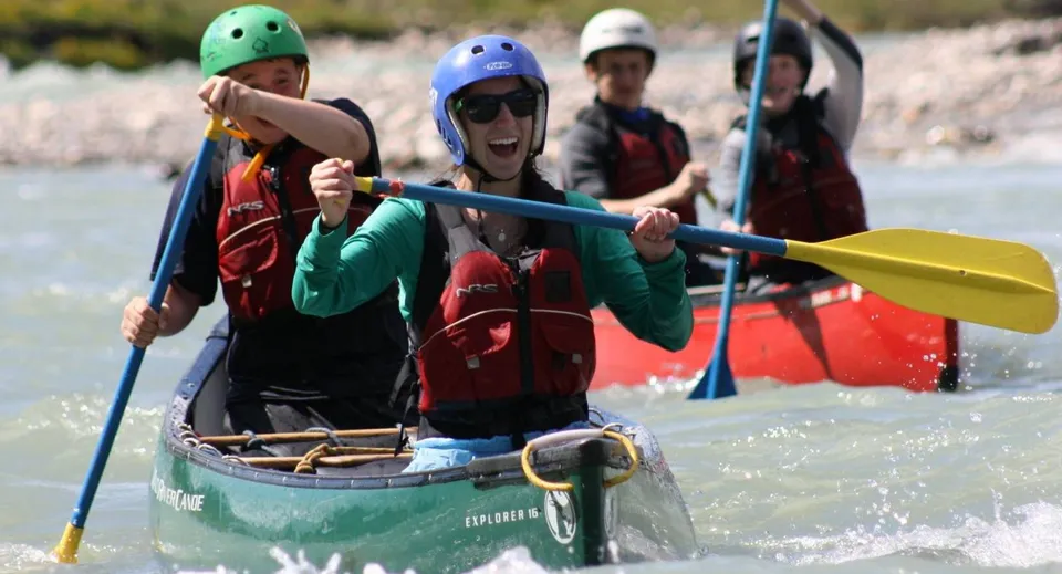 Studenti del Coláiste Bhaile Chláir pagaiano con caschi colorati su una piccola barca verde lungo un fiume attraverso un paesaggio erboso.