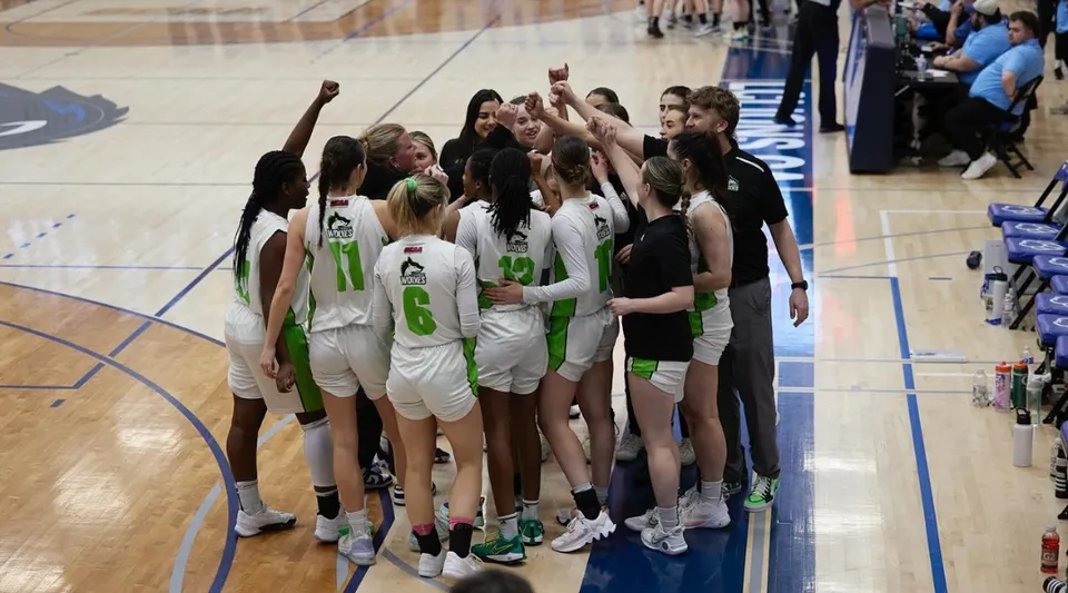Un gruppo di giocatrici di basket in maglia sta sul campo da gioco dell'Algonquin College con gli spettatori sullo sfondo.