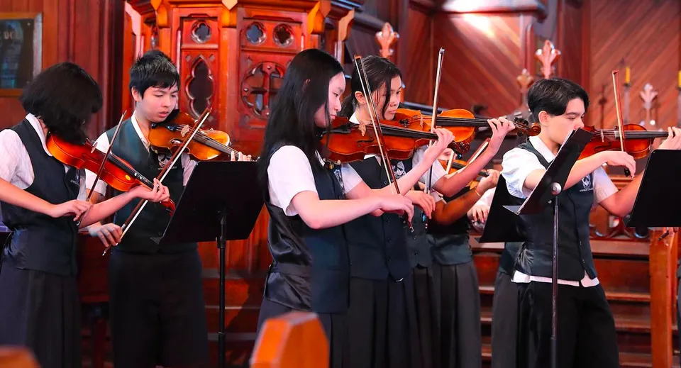 Un gruppo di musicisti in abiti eleganti si esibisce sul palco del Lynfield College davanti a uno sfondo rosso e oro.