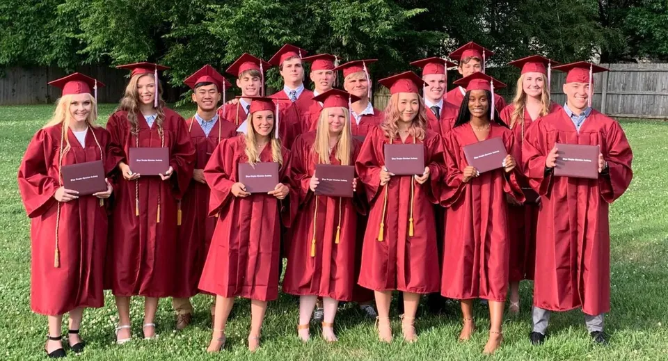 Un gruppo di diplomati della Plaza Heights Christian Academy sta all'aperto davanti ad alberi verdi indossando toghe rosse da diploma.