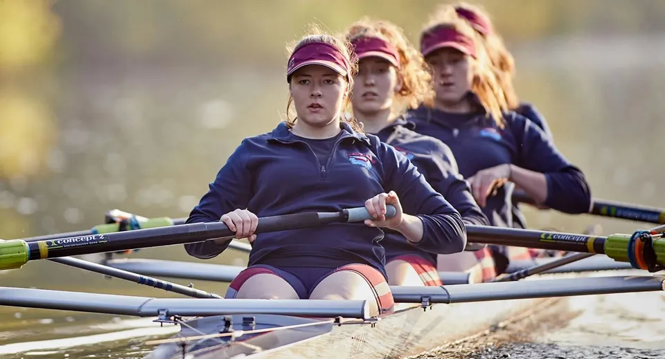 Un gruppo di studentesse della St. James’ School rema su uno specchio d'acqua con un paesaggio naturale sullo sfondo.