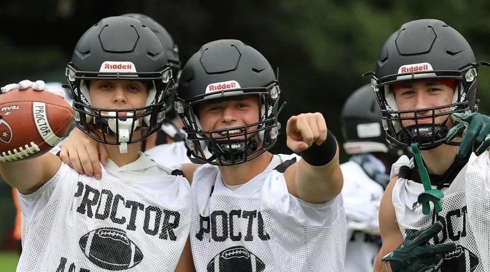 Tre studenti della Proctor Academy in uniforme da football stanno insieme e tengono un pallone da football su un campo sportivo.
