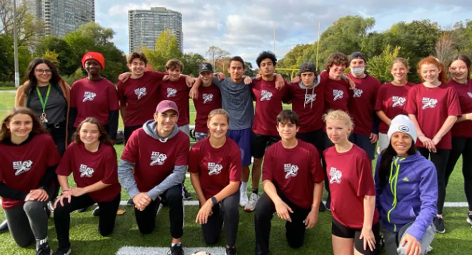 Un gruppo di studenti dell'Etobicoke Collegiate Institute indossa magliette bordeaux coordinate e si trova insieme in un parco davanti a dei grattacieli.