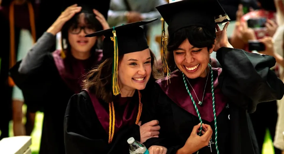 Due diplomate della Idyllwild Arts Academy si abbracciano nelle loro toghe di laurea davanti a un gruppo di altri diplomati.