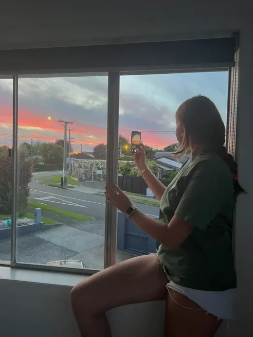 Chica fotografiando con el móvil la puesta de sol desde la ventana de su habitación