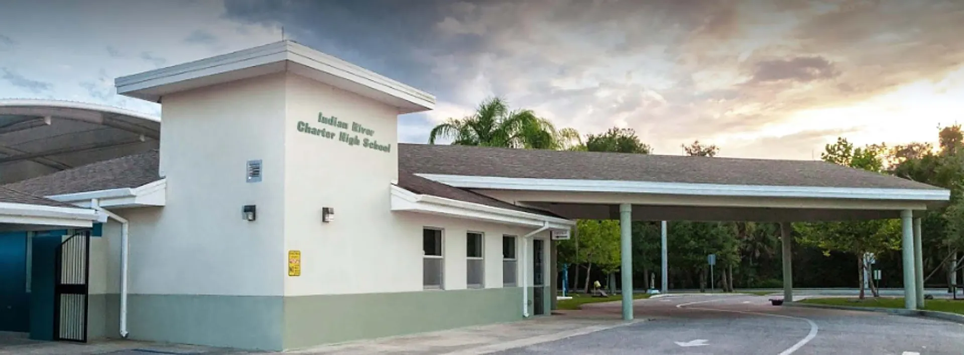 Il moderno edificio bianco e verde della Indian River Charter High School con ingresso coperto e palme sullo sfondo, sotto un cielo nuvoloso.