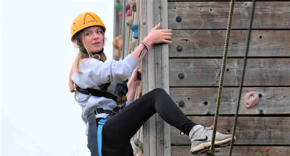 Una studentessa del Thornton College si arrampica su una struttura in legno indossando casco protettivo ed equipaggiamento di sicurezza.