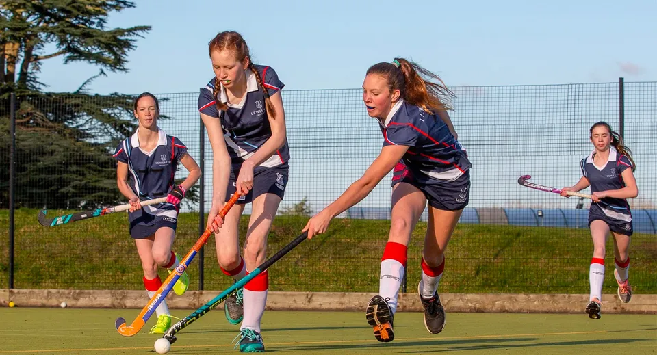 Studentesse della Leweston School giocano a hockey su prato su un campo erboso con alberi e una recinzione sullo sfondo.