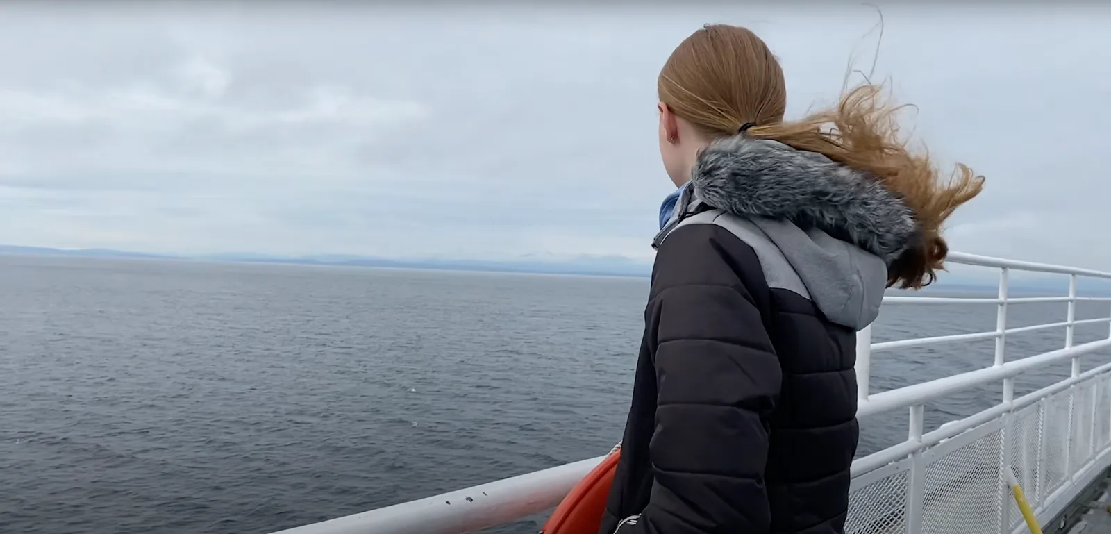 Jugendliche in Winterjacke blickt von der Reling eines Schiffes auf das graue Meer