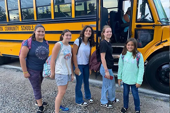 Cinco estudiantes con mochilas posan sonrientes junto a un autobús escolar amarillo