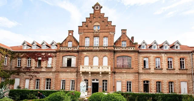 Das Gebäude des Lycée Notre Dame de Bon Secours mit einem markanten Treppengiebel und Bogenfenstern ist von üppigem Grün umgeben.