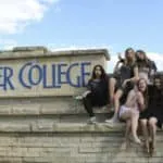 Varias estudiantes sonrientes sentadas en un muro de piedra junto a un cartel universitario