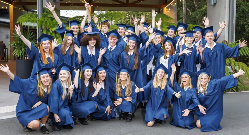 Un grande gruppo di diplomati festeggia in toga e tocco blu sul campus della Coolum State High School.