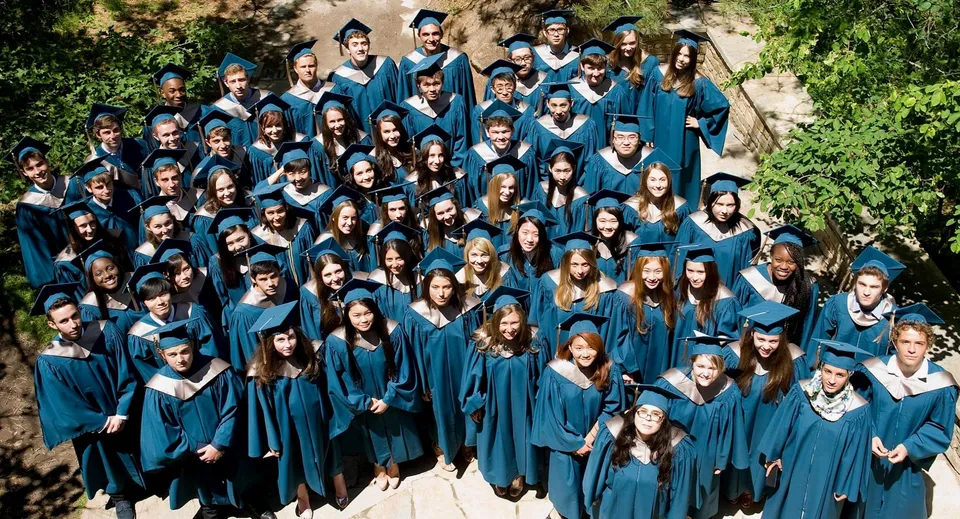 Un grande gruppo di diplomati della Ross School si trova all'aperto con toghe e tocchi blu davanti ad alberi e aree verdi.