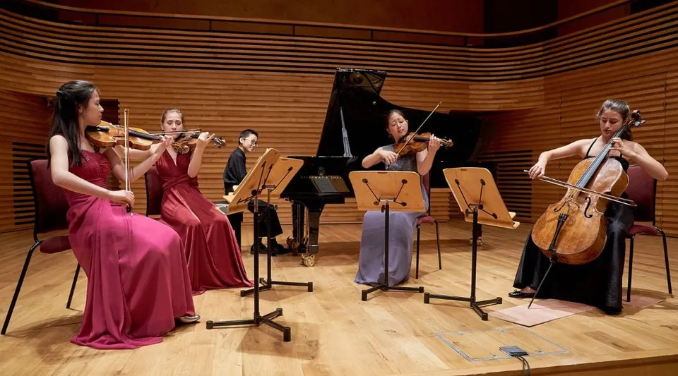 Un gruppo di studenti di musica della Yehudi Menuhin School si esibisce in abiti eleganti su un palco con un pianoforte a coda e pareti in legno.